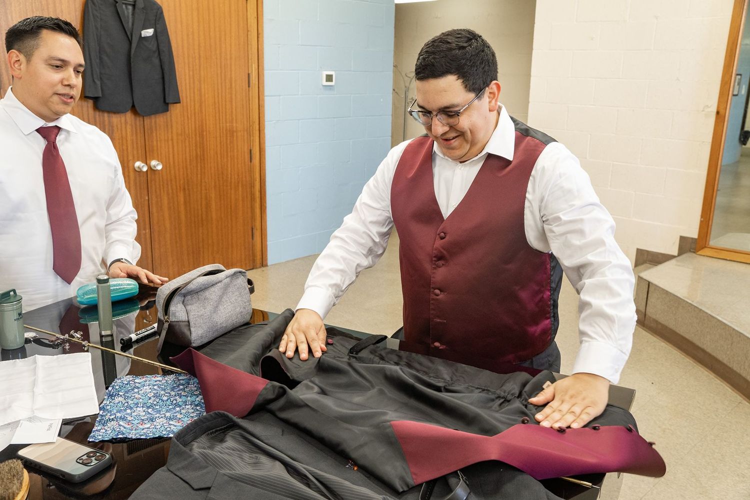 Groom in burgundy vest arranges his wedding day accessories on a table before the ceremony.