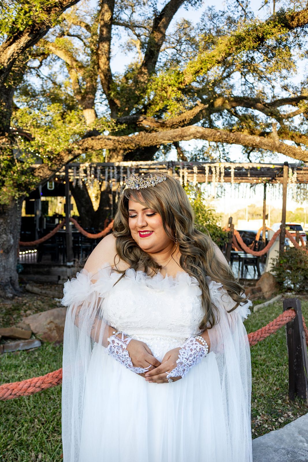 Elegant bride wearing a white gown, tiara, and lace gloves posing outdoors under a large oak tree.