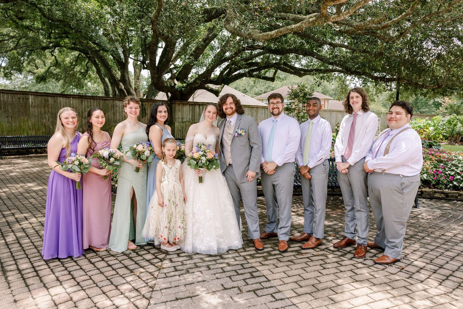 Wedding party poses under large oak trees wearing purple, blue and gray attire.