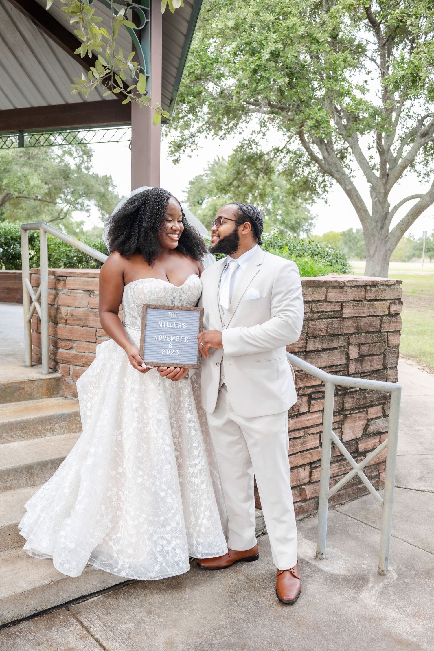 A couple in wedding attire share a joyful moment while holding a sign on stone steps under a gazebo.
