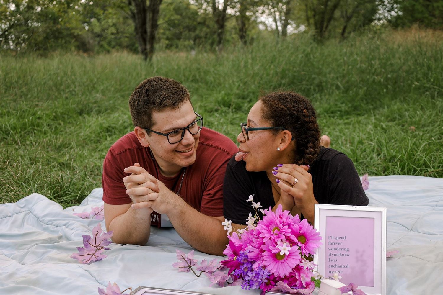 Couple sharing a romantic picnic on a blanket with pink flowers, gazing at each other in a lush green field.