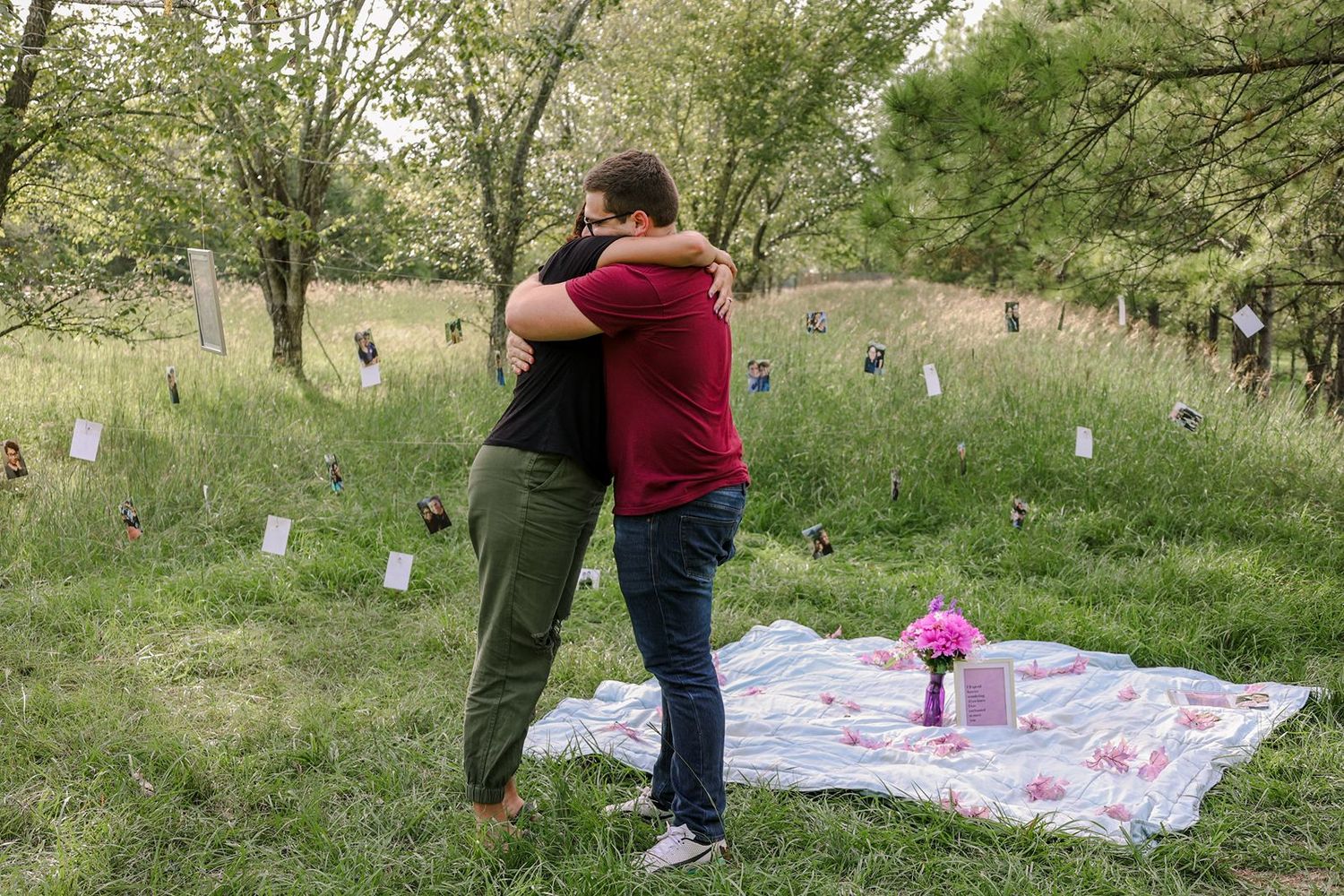 Two people share an emotional hug in a field memorial with white markers and pink flowers on a blanket.
