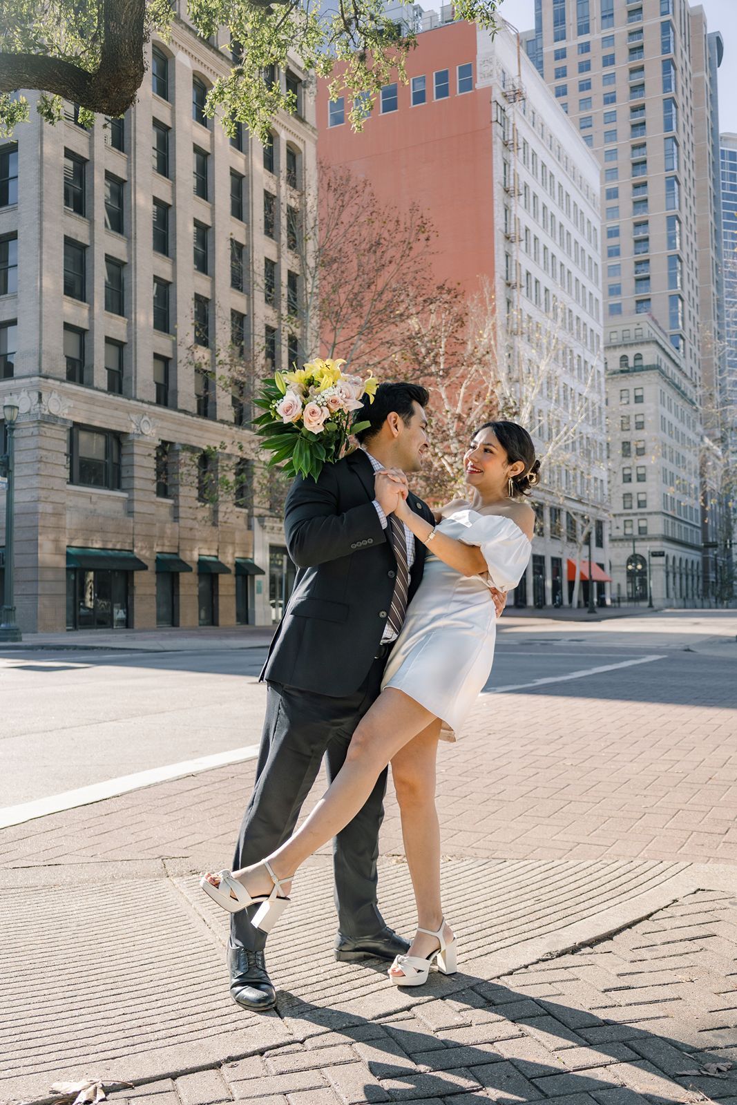 A couple dances together on a sunlit city street with tall buildings in the background.