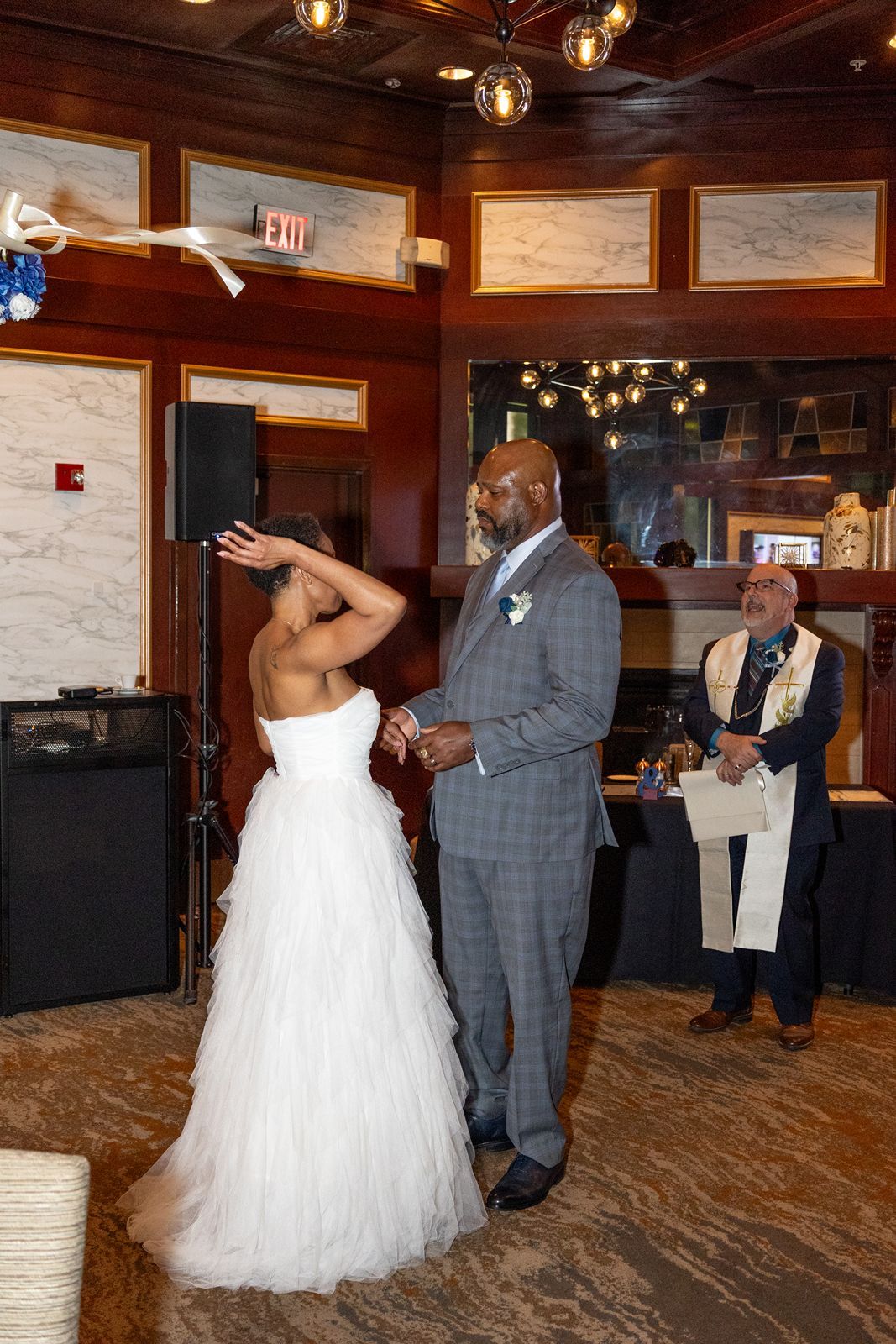 Bride and groom share first dance at wedding reception in elegant wood-paneled venue with officiant watching nearby.
