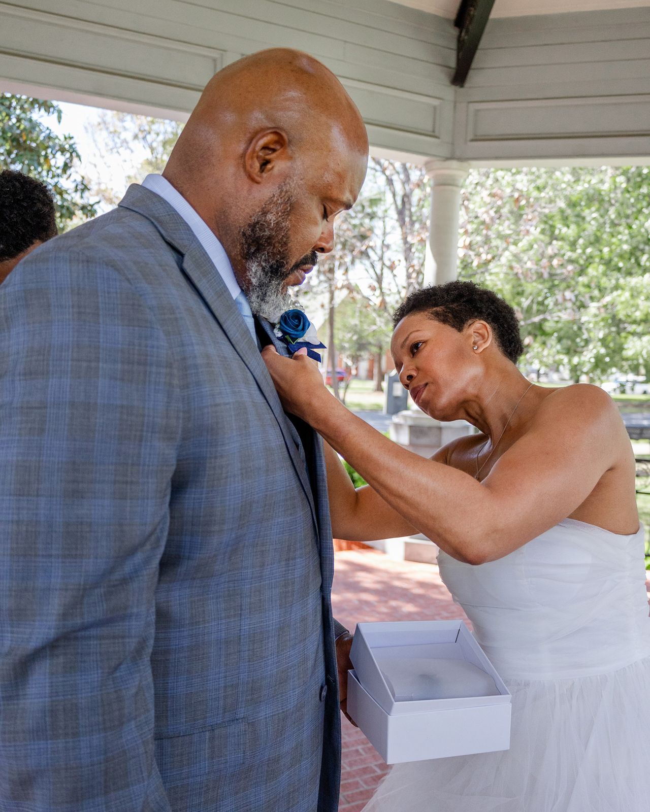 Bride in strapless white gown carefully pins blue boutonniere onto groom's gray plaid suit lapel.