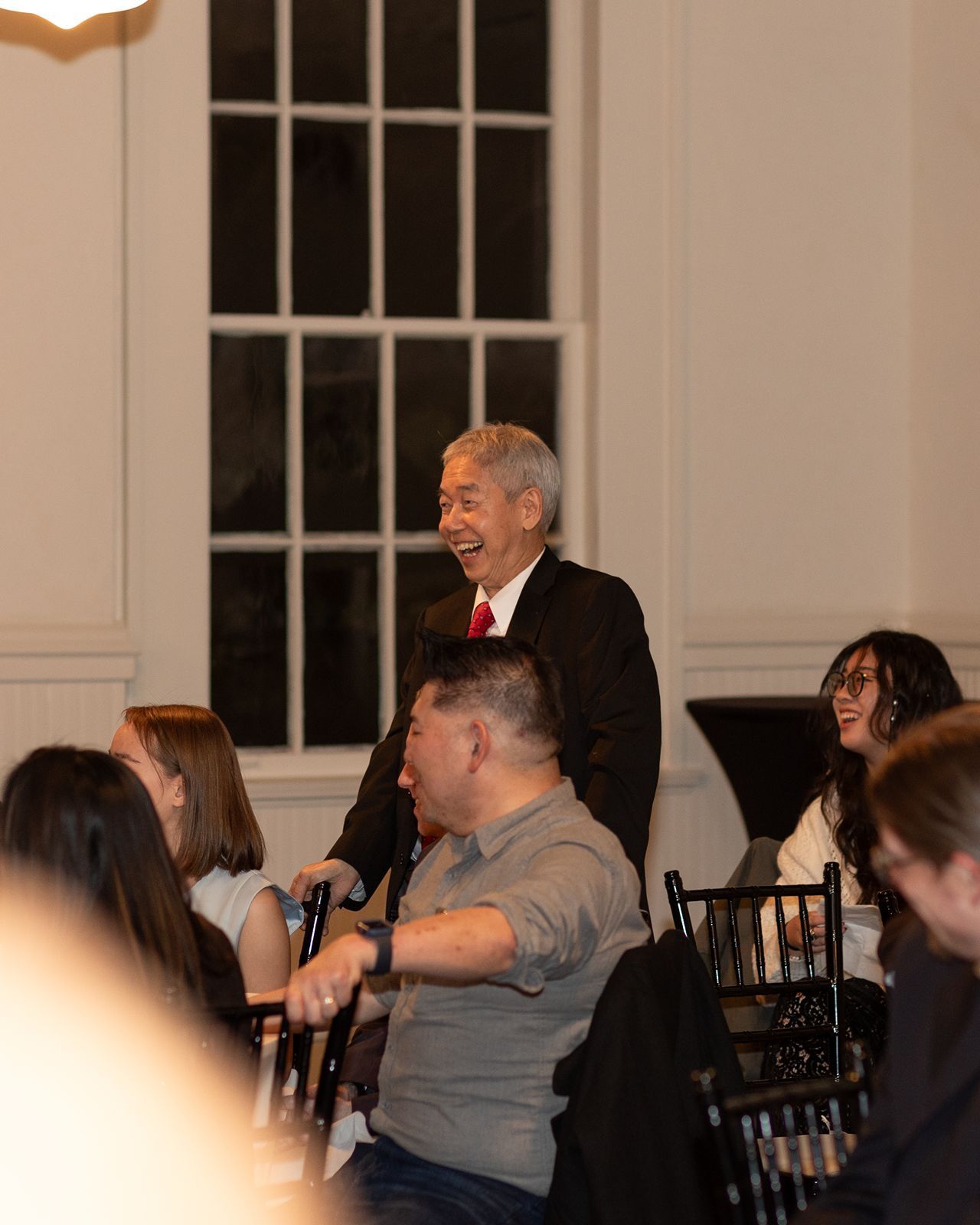 People seated in wooden chairs listen attentively during an indoor event in a room with large windows.
