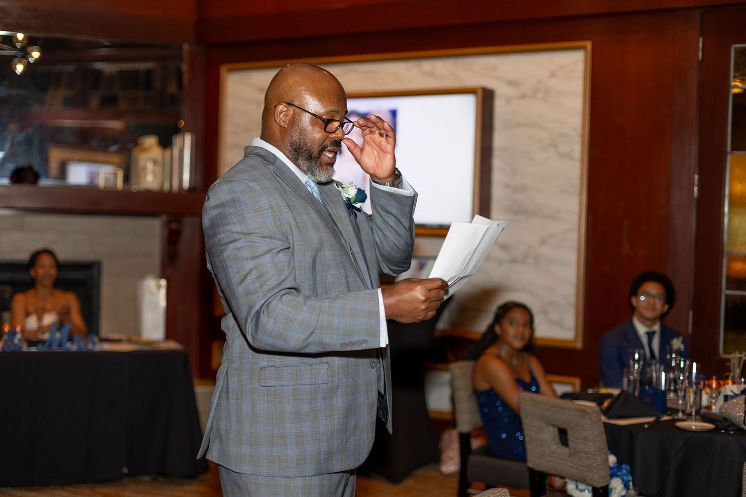 Man in gray plaid suit reading from papers while speaking at a formal wedding event.