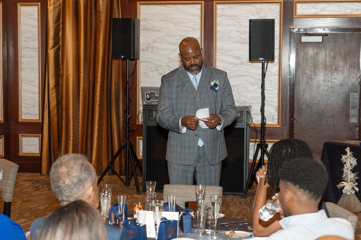 A man in a gray suit delivers a speech at an elegant wedding reception dinner with guests seated at decorated tables.