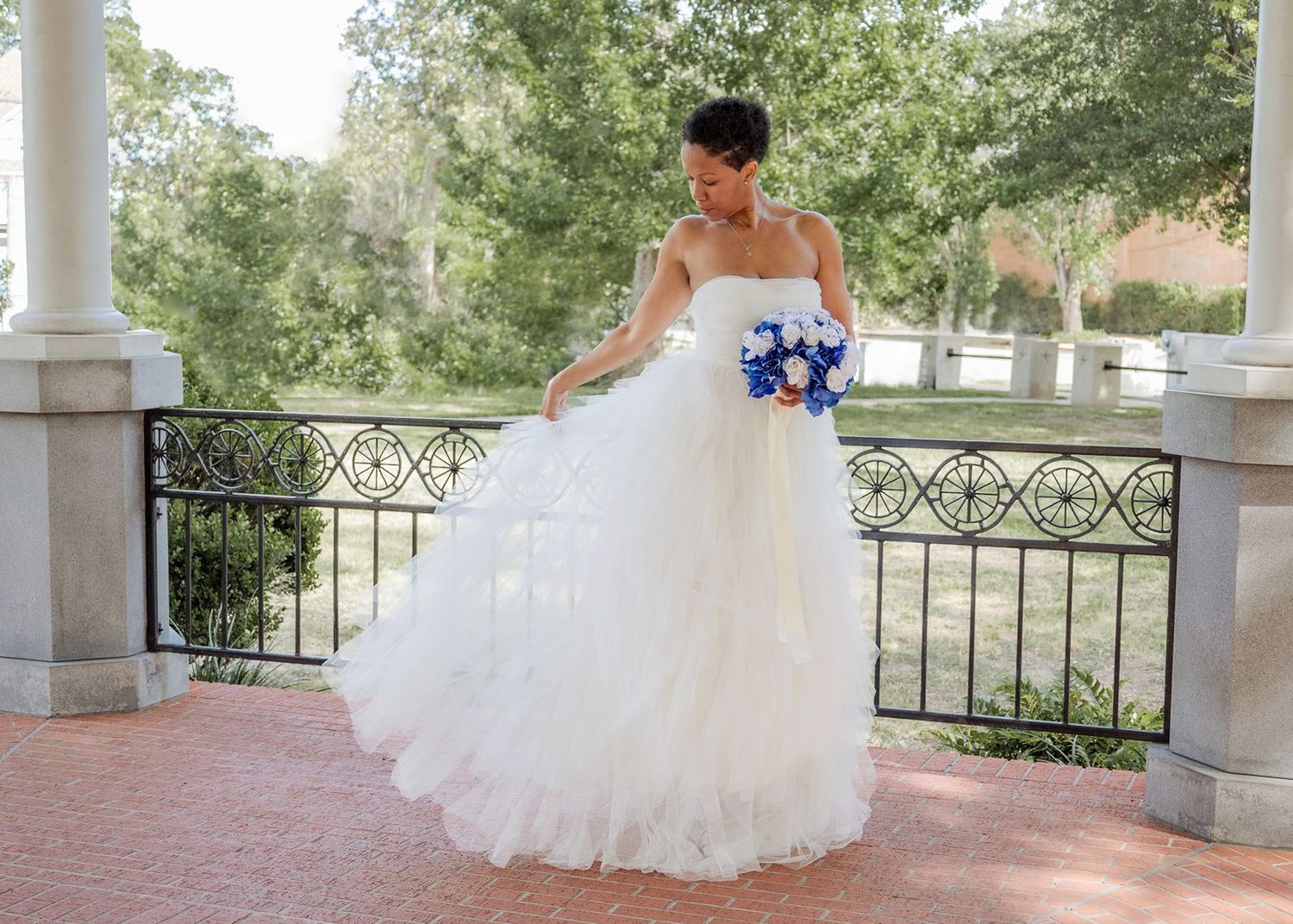 Bride in strapless white ballgown holding blue bouquet on elegant outdoor venue with columns.