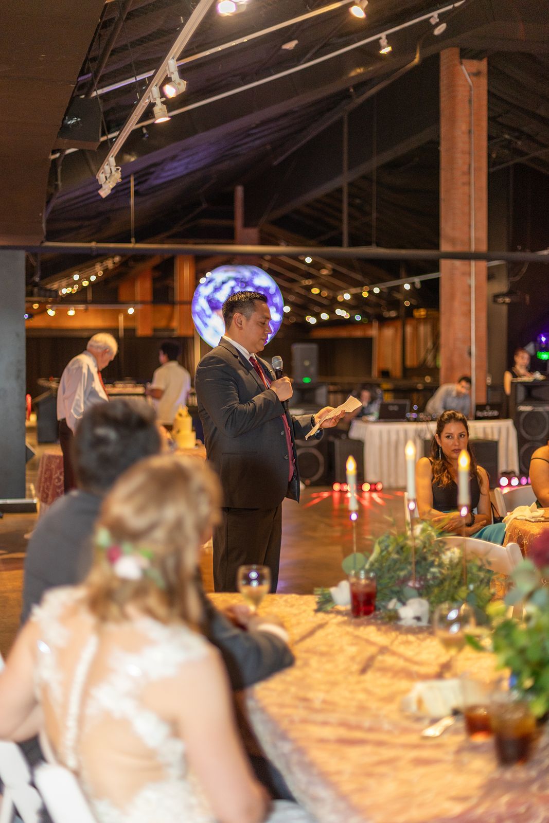 A man in a suit gives a speech at an elegant wedding reception in a rustic venue with string lights.
