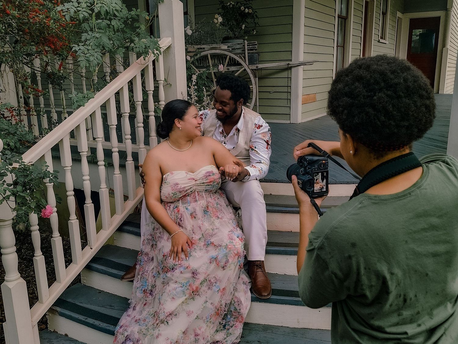 Photographer captures couple posing on porch steps, woman in floral dress and man in white suit, during outdoor photo shoot.