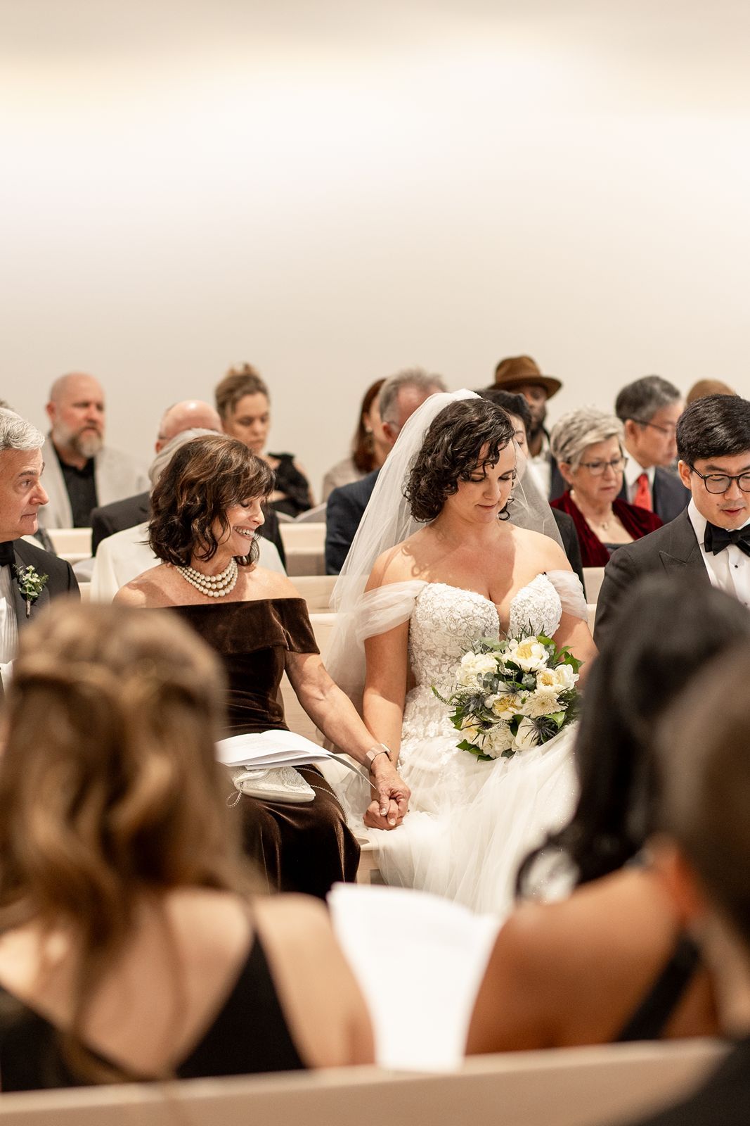 Wedding ceremony in progress with attendees seated in pews as bride in off-shoulder white dress holds bouquet of white flowers.