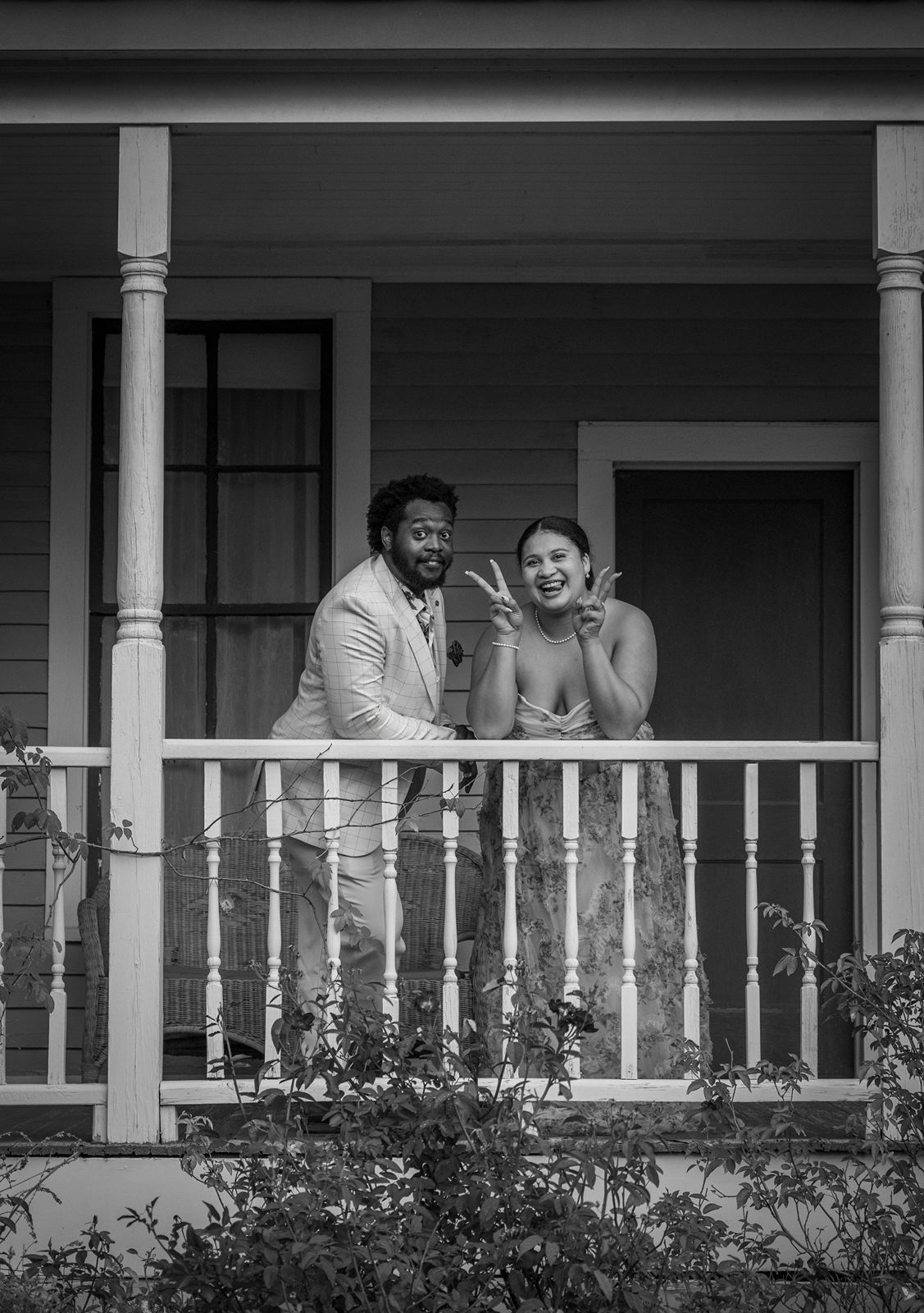 A couple shares a joyful moment together on a white porch with decorative columns and flowering bushes below.