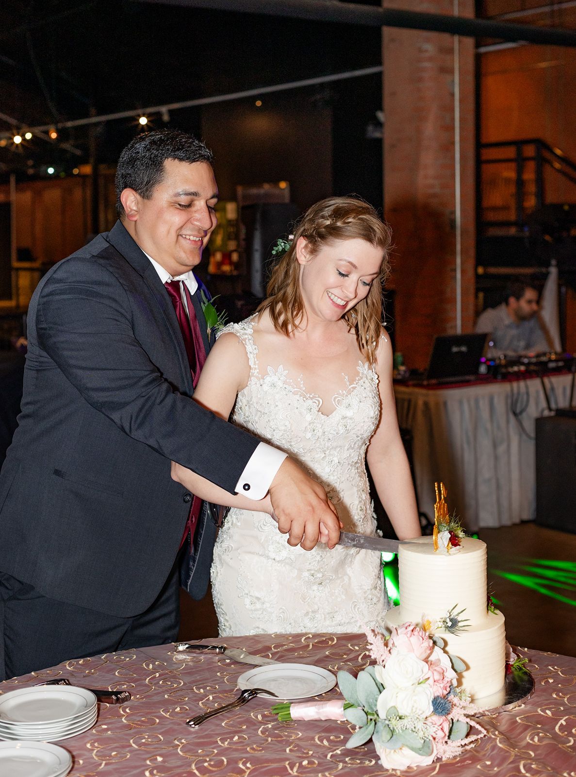 A couple cuts their wedding cake together at an indoor reception venue with brick walls and ambient lighting.