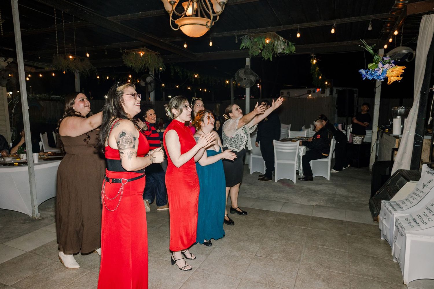 Women in elegant dresses gather at an outdoor evening wedding reception, watching the bouquet toss under string lights.