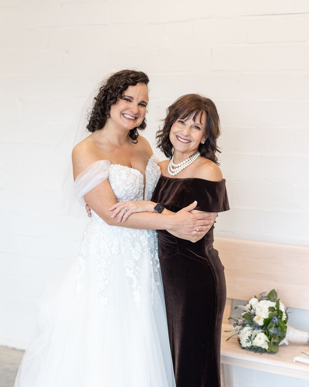 Two people share a joyful embrace in a bright room while wearing a white wedding dress and black formal attire.