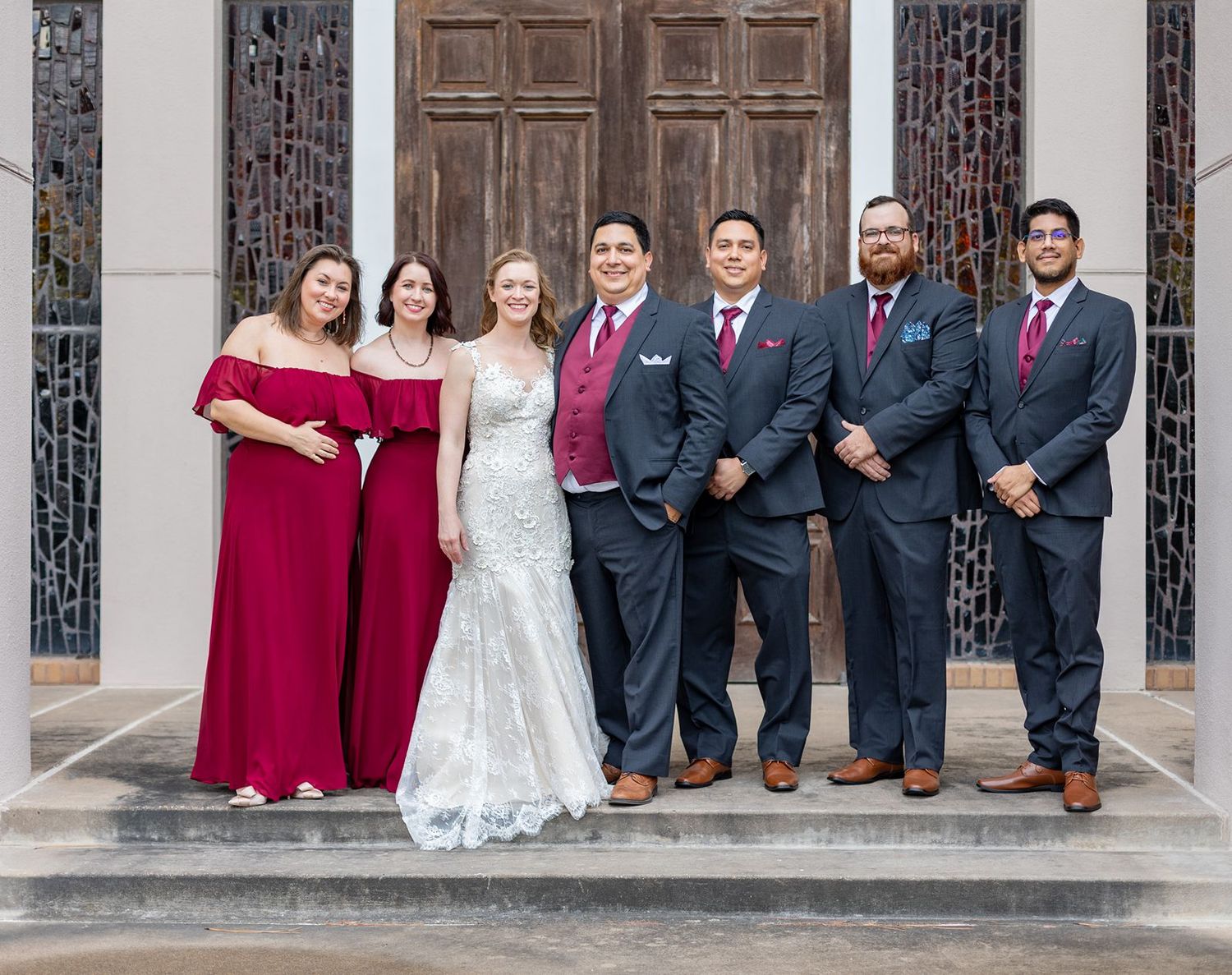 Wedding party in formal attire poses together on stone church steps with dark wooden doors and ornate details.