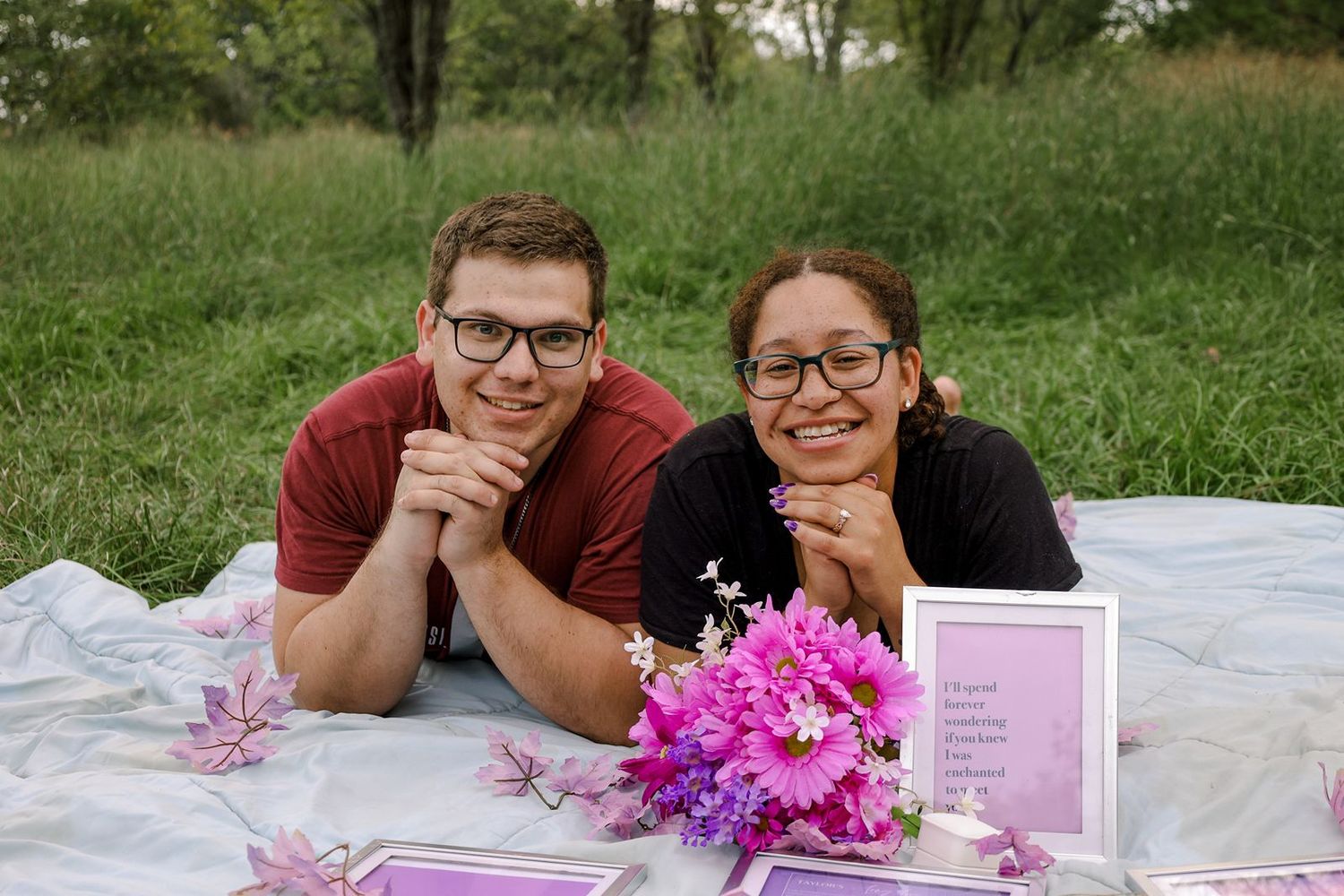 Couple smiling on a white blanket outdoors with pink flowers and framed cards in a grassy field.