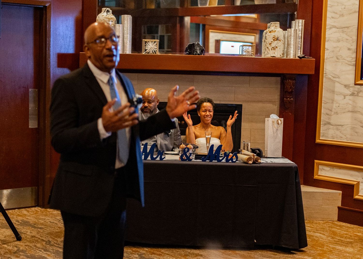Man gestures while speaking at wedding reception as bride claps joyfully in background.