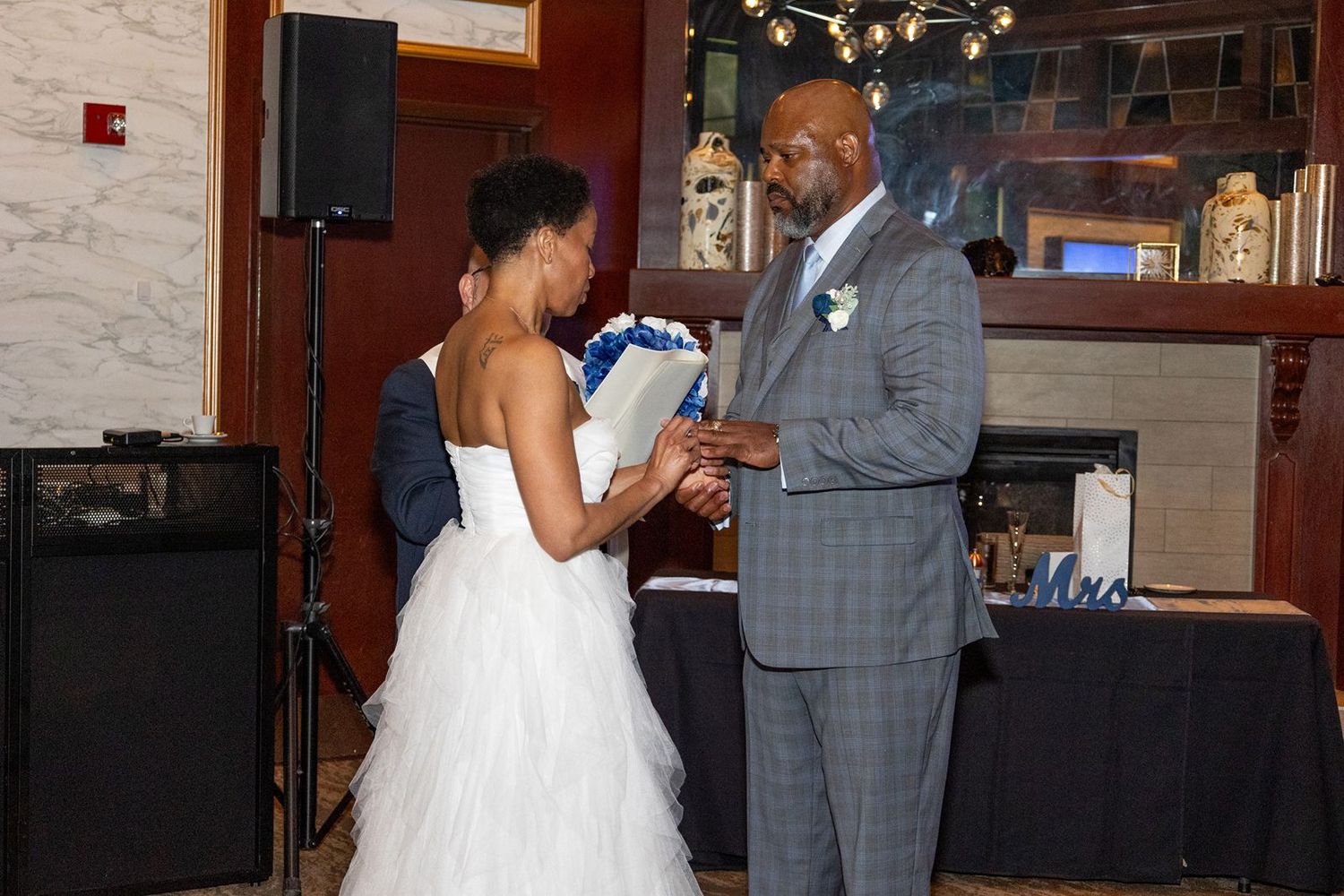 Couple exchanges vows at indoor wedding, bride in white gown holds blue bouquet.