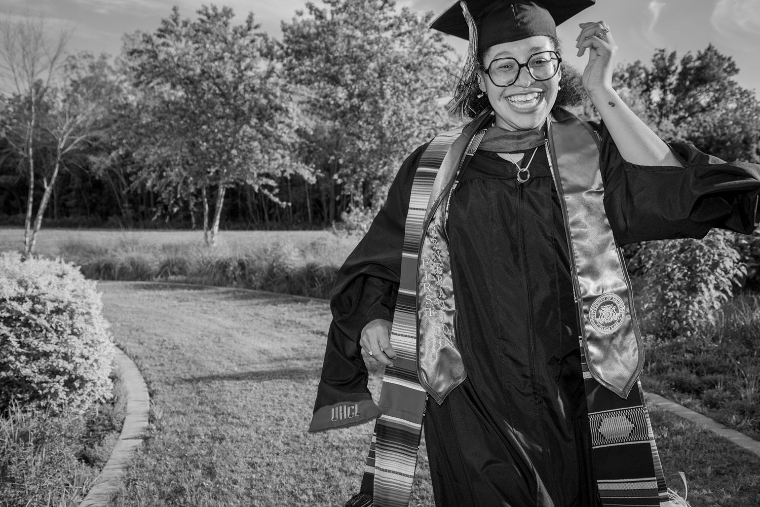 Happy graduate in black cap and gown adjusts mortarboard while smiling outdoors in black and white photo.