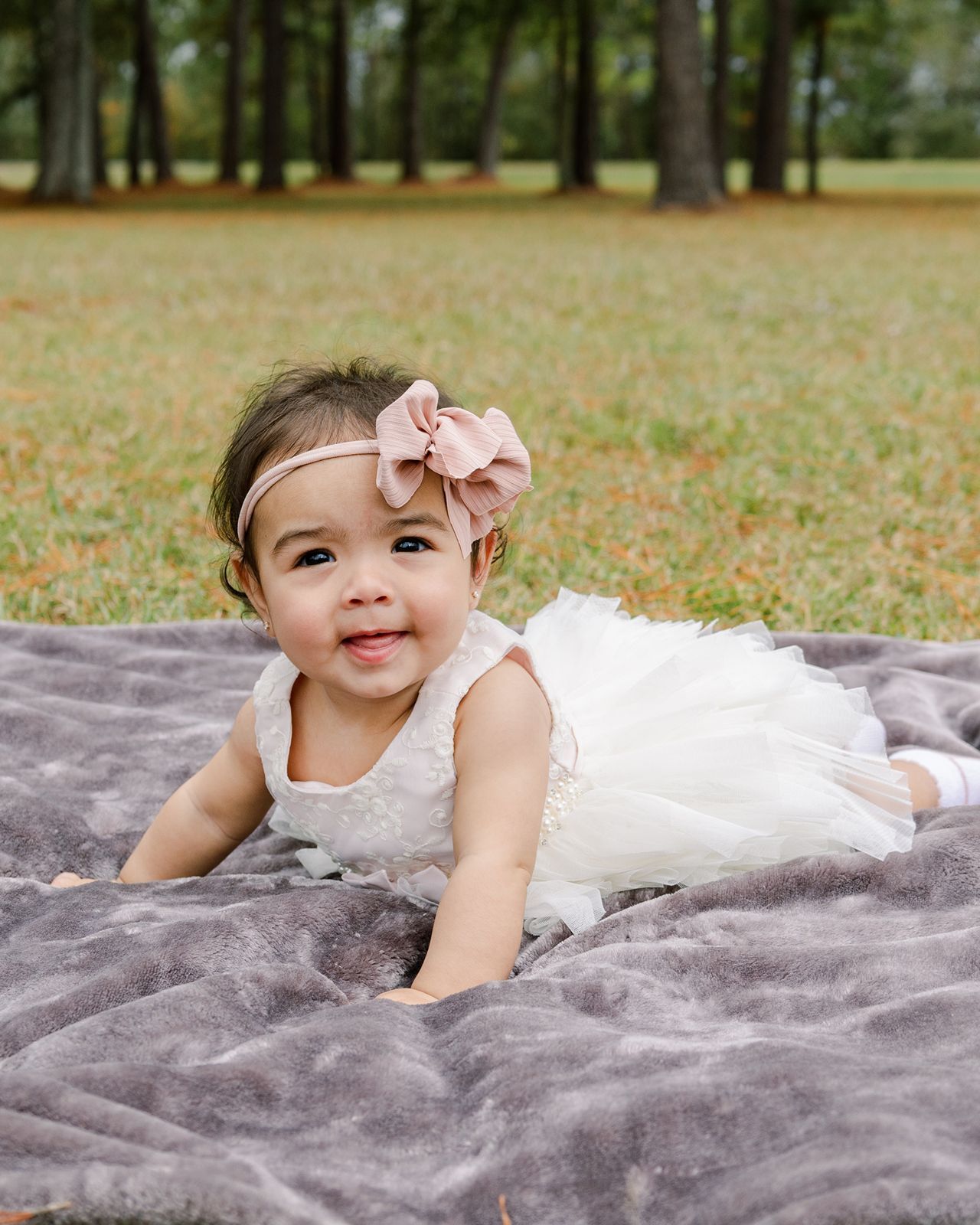 Small child in white dress lying on gray blanket outdoors with autumn trees in background.