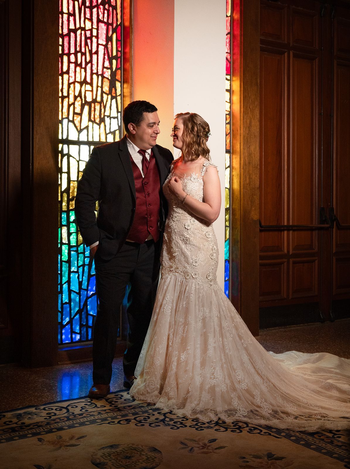 Bride and groom pose together in front of colorful stained glass windows during their wedding ceremony.