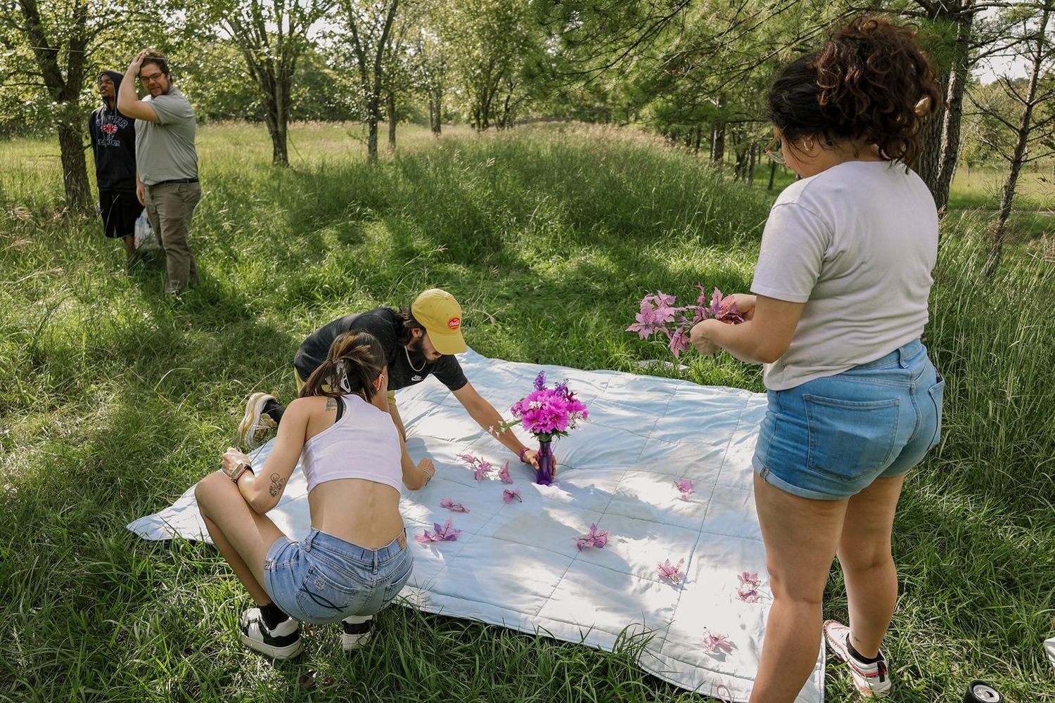 Two young women arranging flowers on a white blanket in a lush green meadow during a picnic setup.