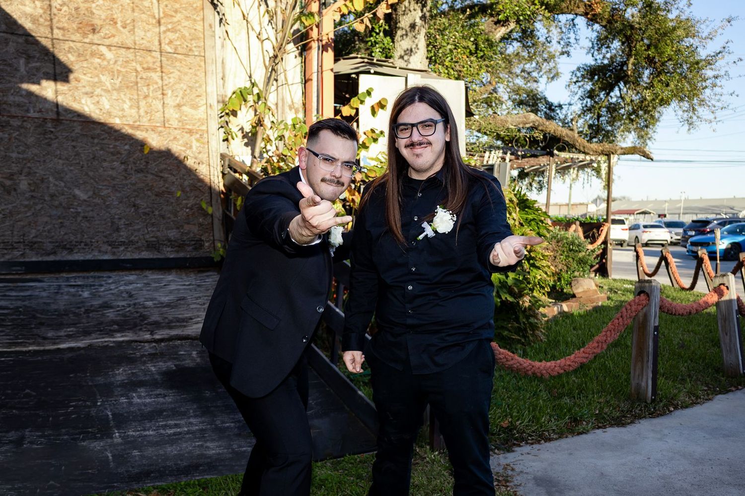 Two men in black suits smile and pose outdoors at a wedding venue with greenery and string lights.