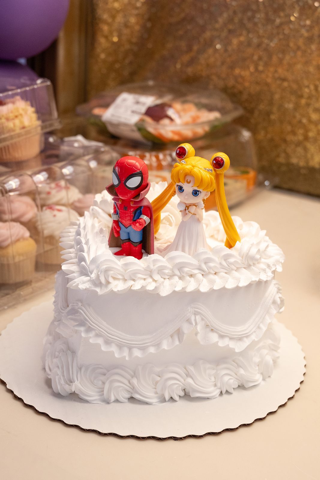 White wedding cake topped with Spiderman and Sailor Moon figurines, decorated with white frosting swirls.