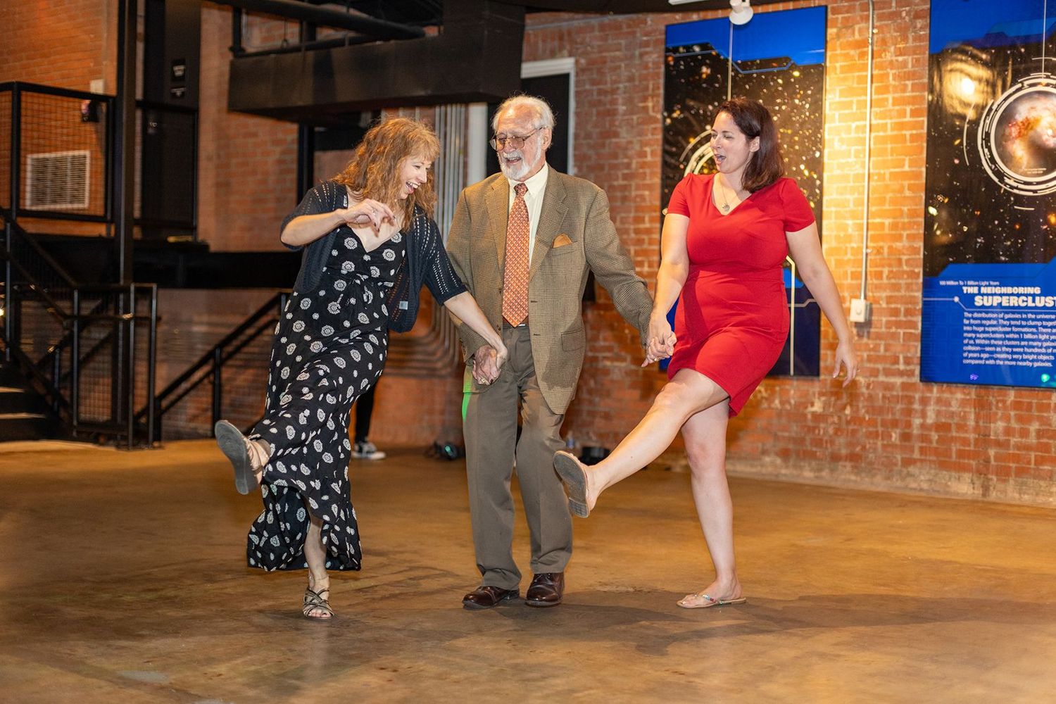 Three people dancing together at an indoor event venue with brick walls.