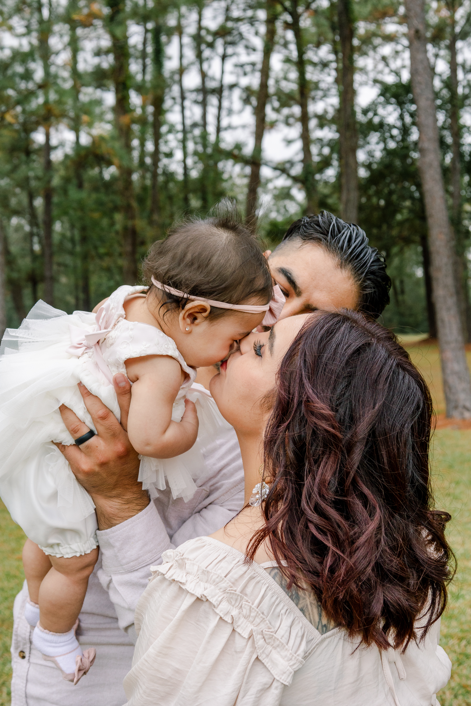 A family shares a tender moment together outdoors in a wooded setting wearing light colored clothing.