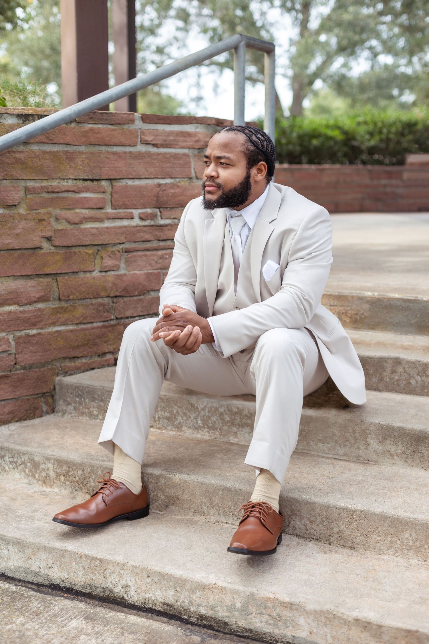 A person in a cream-colored suit sits thoughtfully on concrete steps near a brick wall.