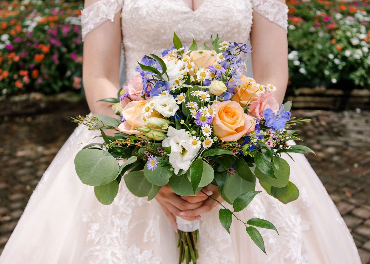 A wedding bouquet featuring orange roses, purple flowers, and green foliage held against a white dress.