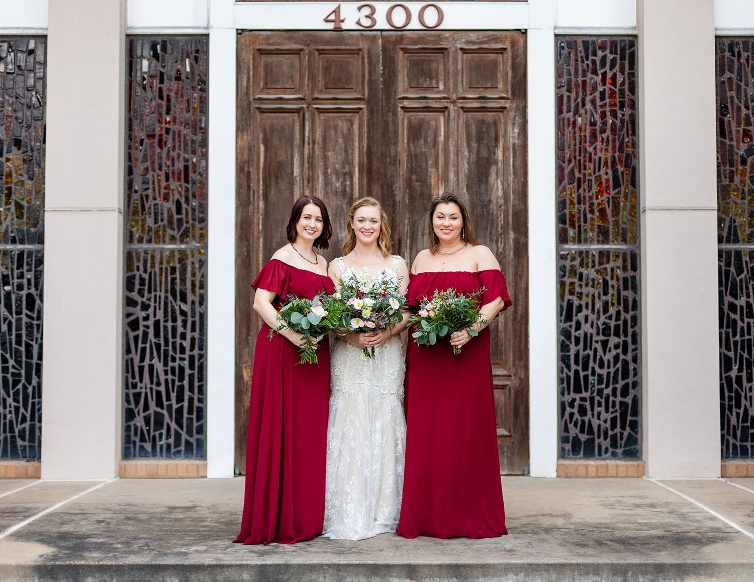 Bride with two bridesmaids in red off-shoulder gowns holding bouquets before church doors numbered 4300.