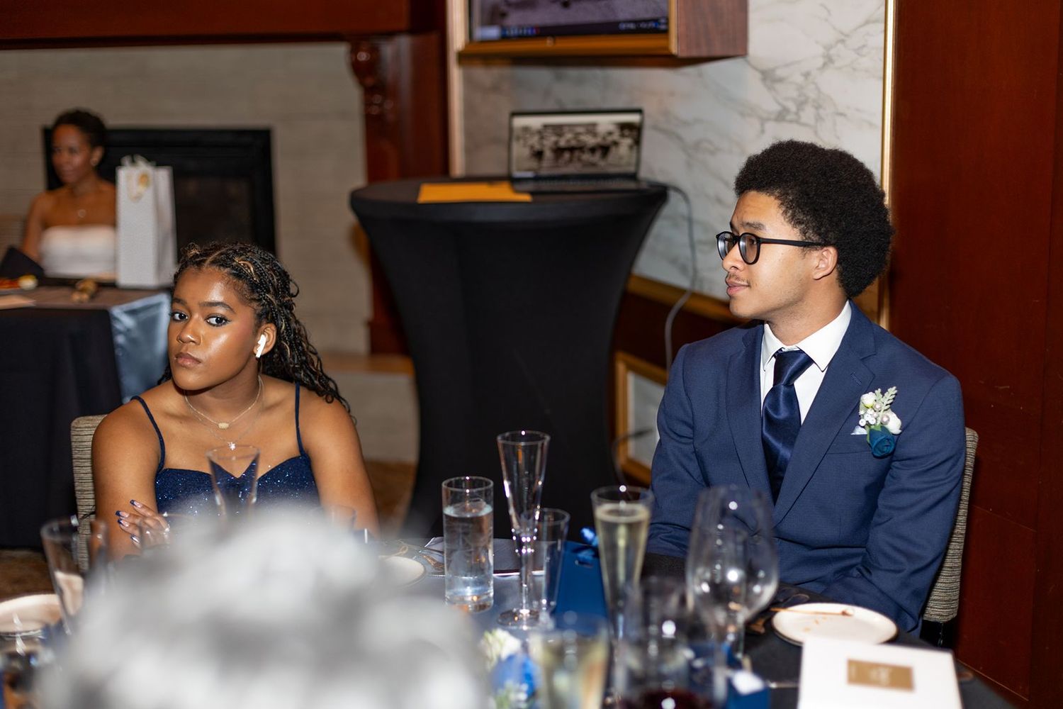 Young couple in formal attire seated at a wedding reception table listening attentively.