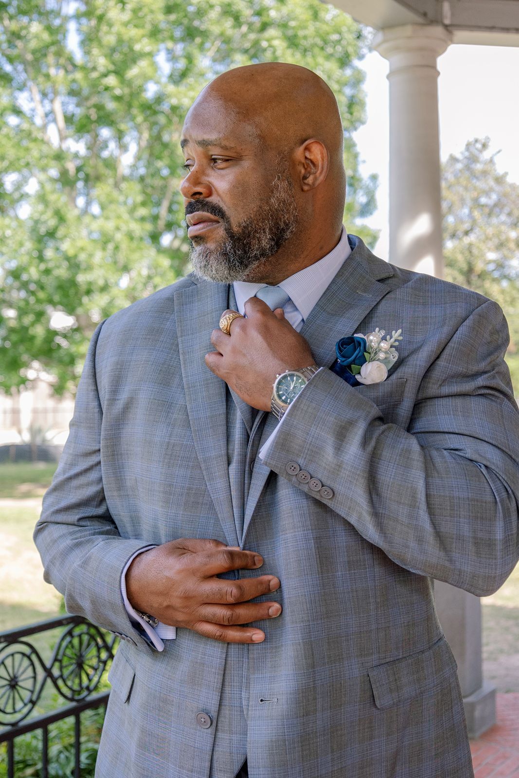 Bald groom in gray plaid suit adjusts tie, wearing boutonniere and watch, standing on outdoor venue porch.