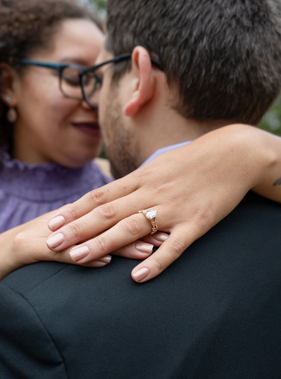 Close-up of a woman's hand displaying a diamond engagement ring resting on a man's shoulder as they embrace.