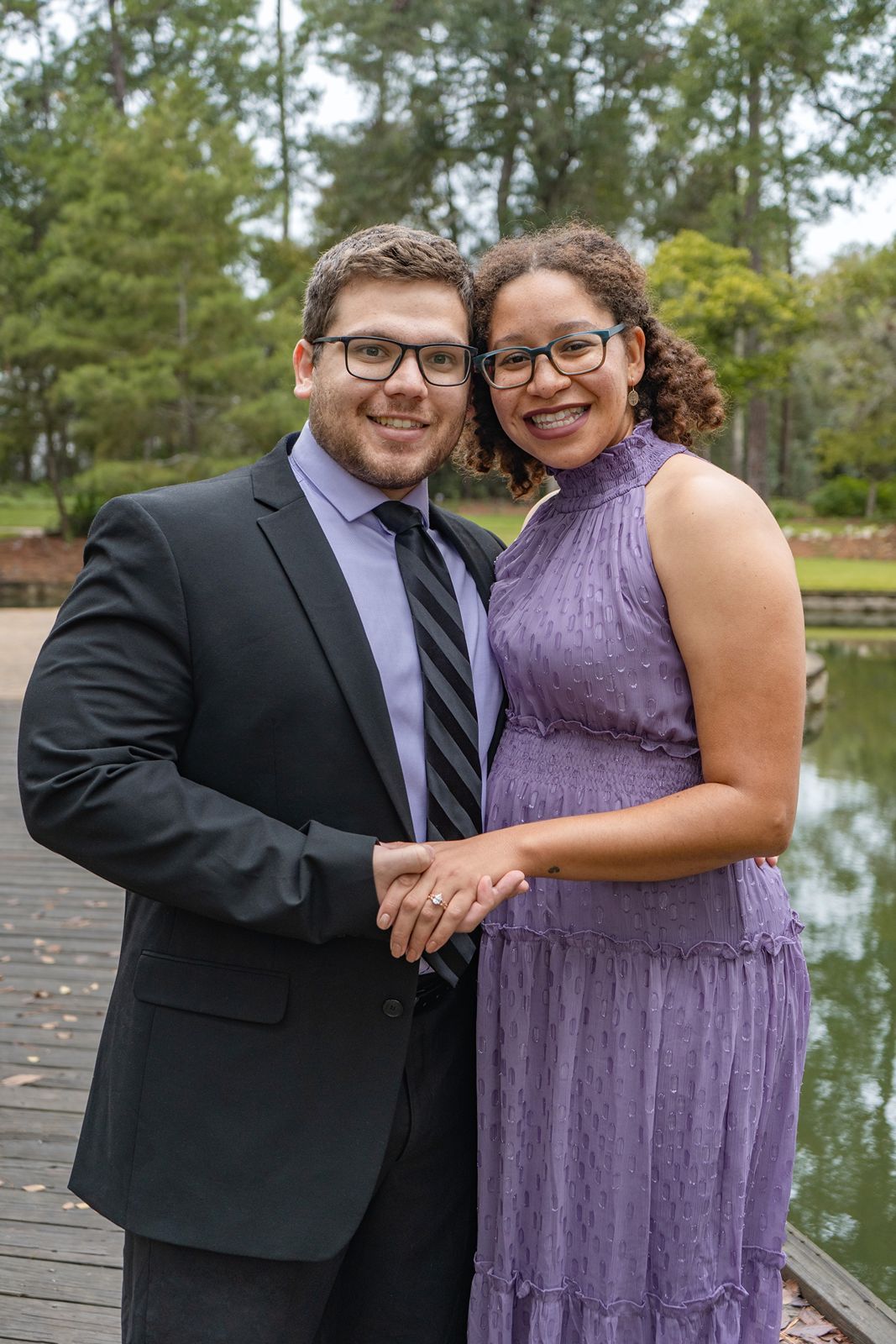 Smiling couple poses on a wooden dock, man in suit and tie, woman in purple dress, holding hands.