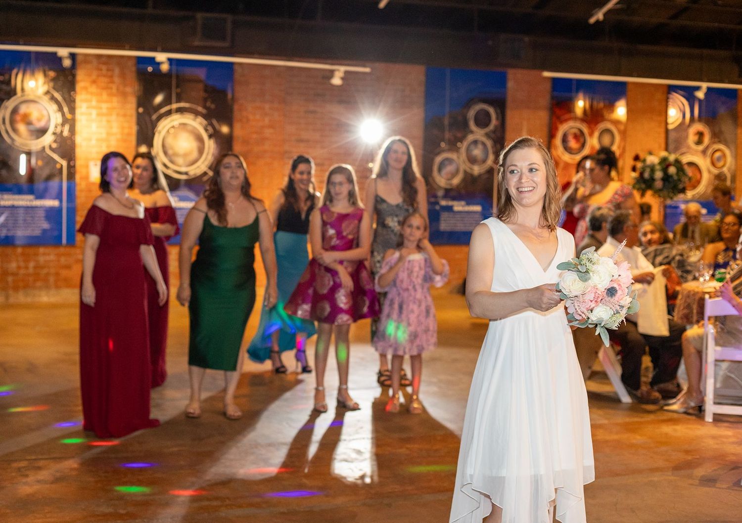 Bride in white dress prepares to toss bouquet to excited bridesmaids in colorful gowns at elegant wedding reception.