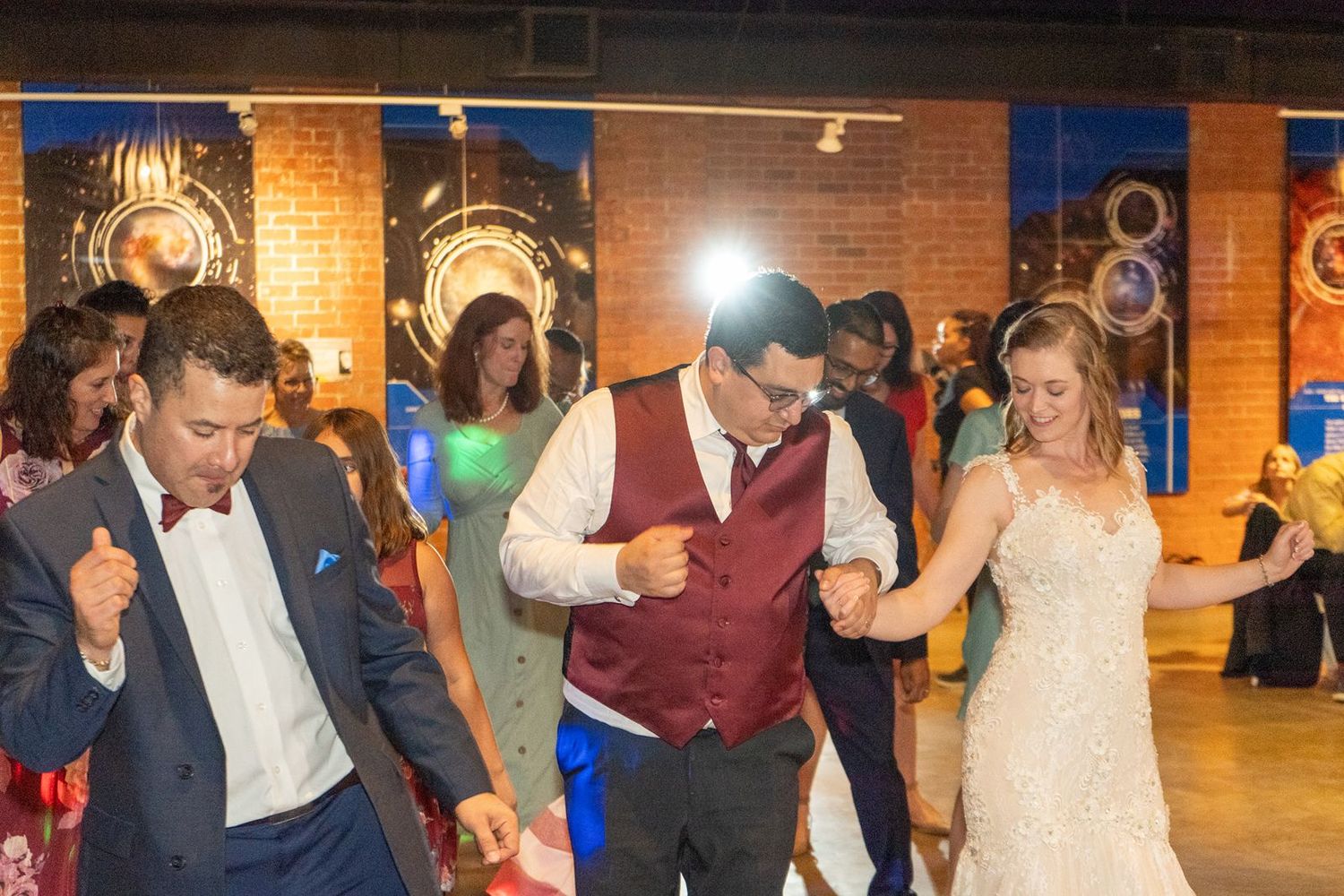 Guests dance joyfully at a wedding reception, including the bride in a white gown and groom in a burgundy vest.