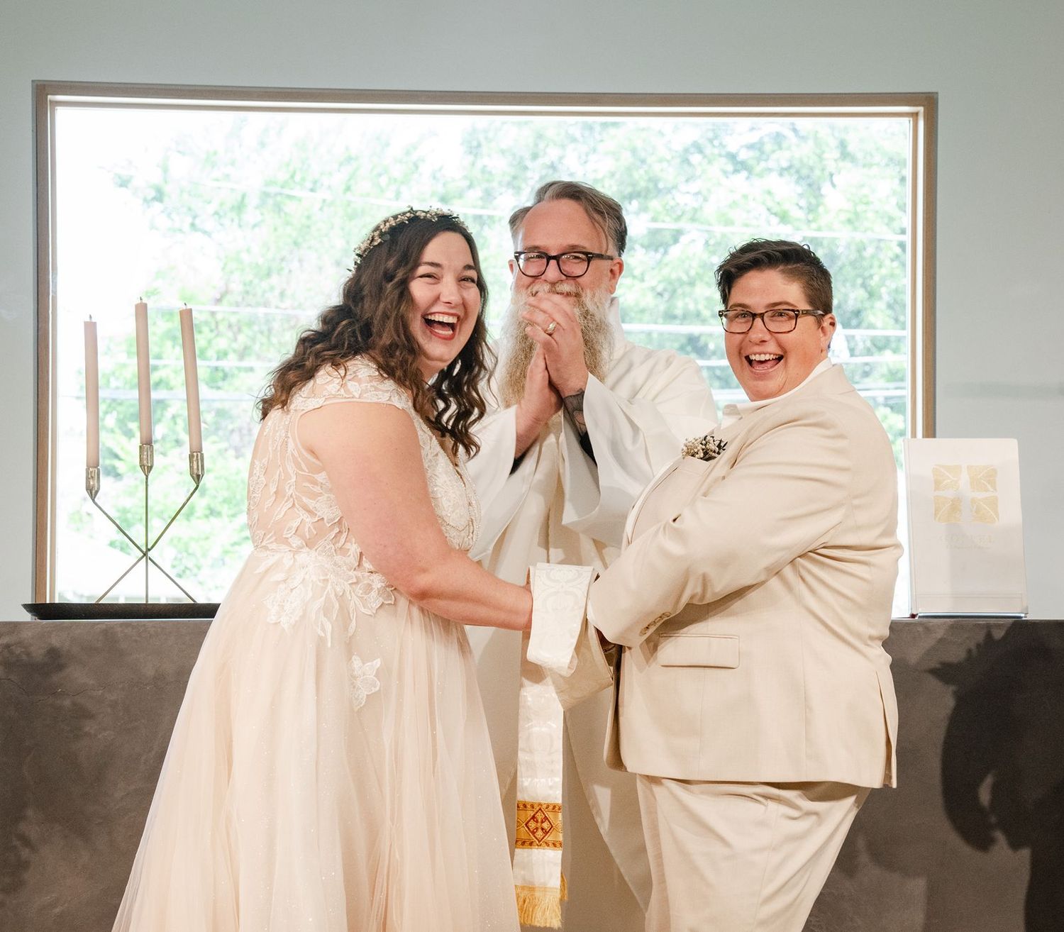 Joyful wedding couple and officiant laugh together during indoor ceremony with candles.