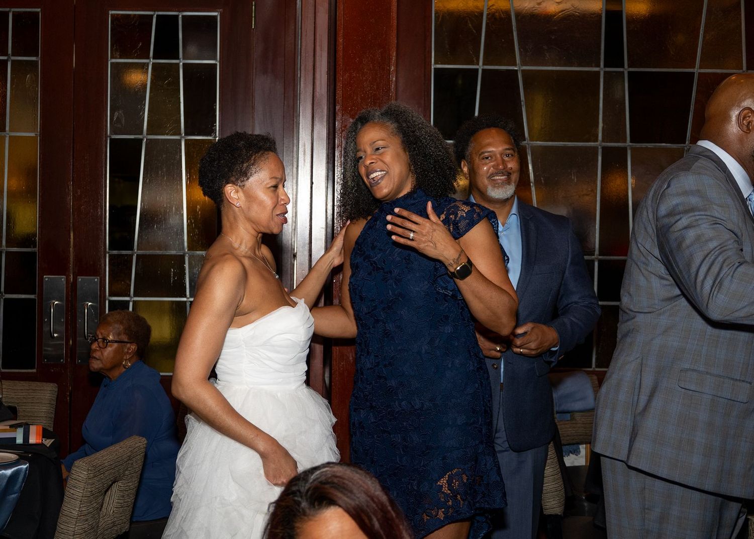 Joyful women in elegant attire embracing at a formal evening event, with guests socializing in background.