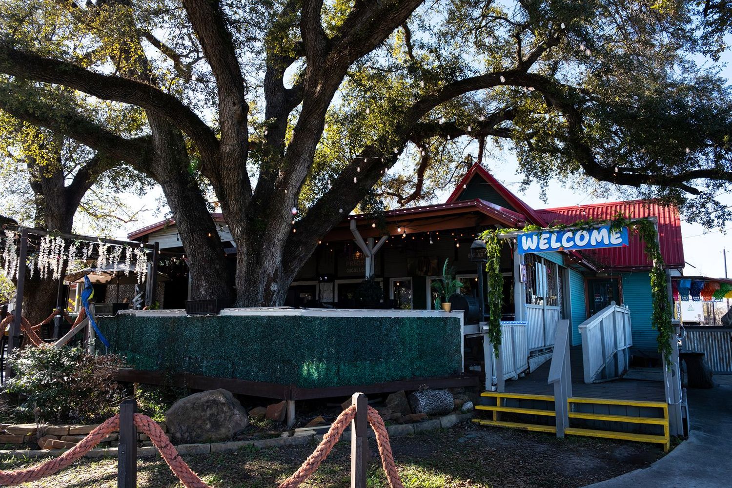 Rustic outdoor restaurant entrance with a large oak tree, colorful welcome sign, and rope fence decor.
