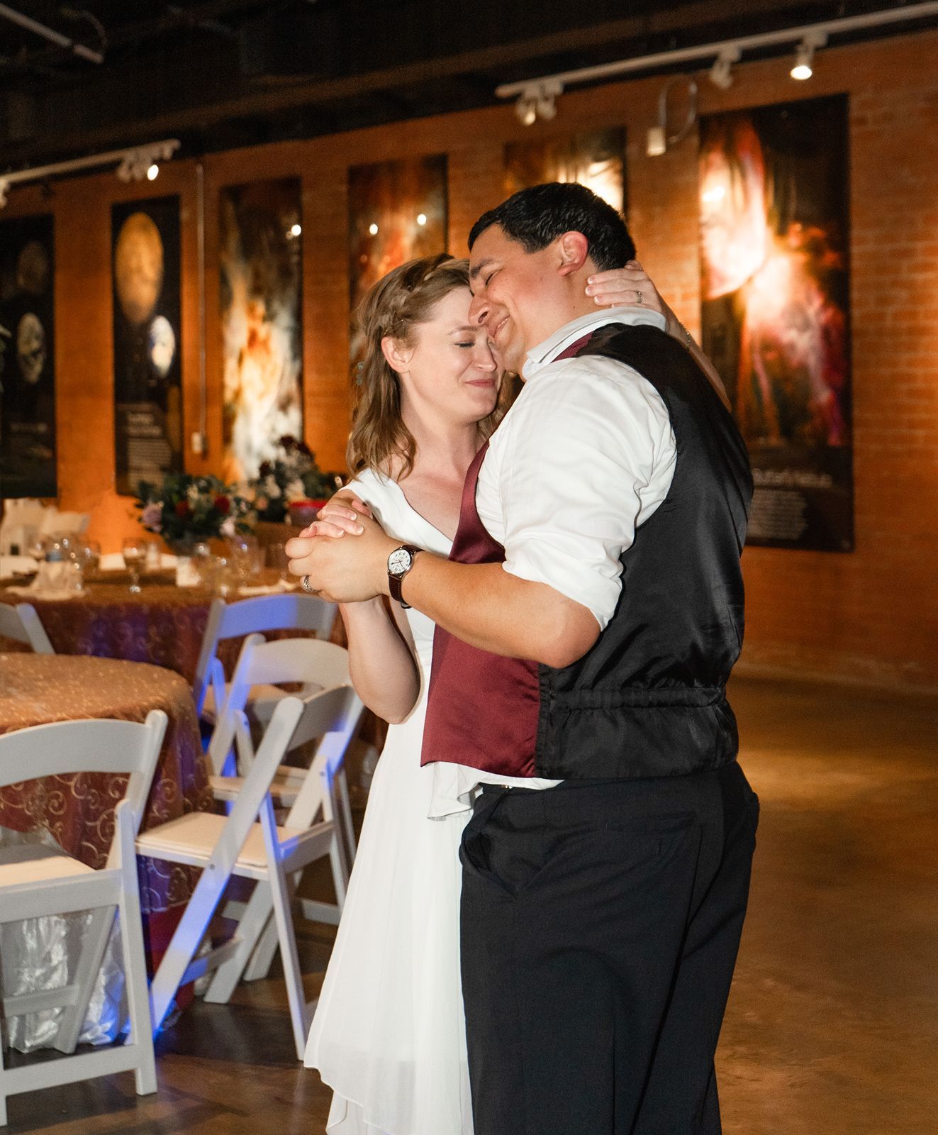 Newlyweds share first dance in a warmly lit reception venue with brick walls.