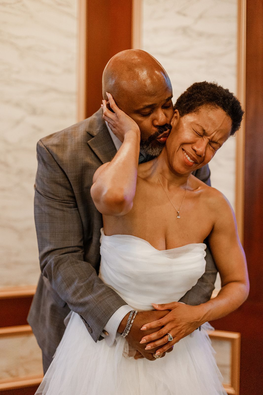 Emotional bride in strapless white gown embraced by groom in gray suit sharing tender wedding moment indoors.