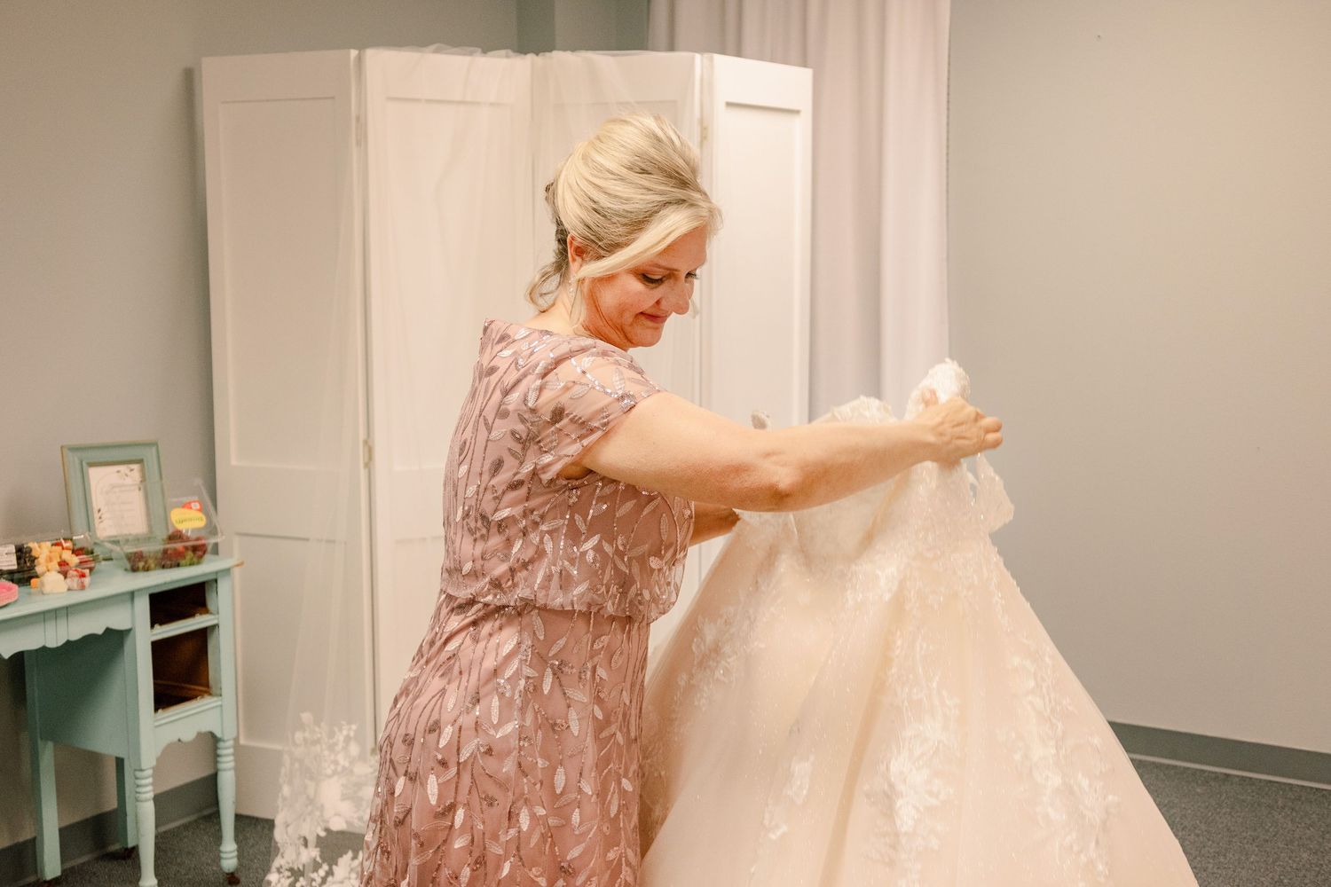 Women in blush pink dress helps woman in white wedding gown with final dress preparations.