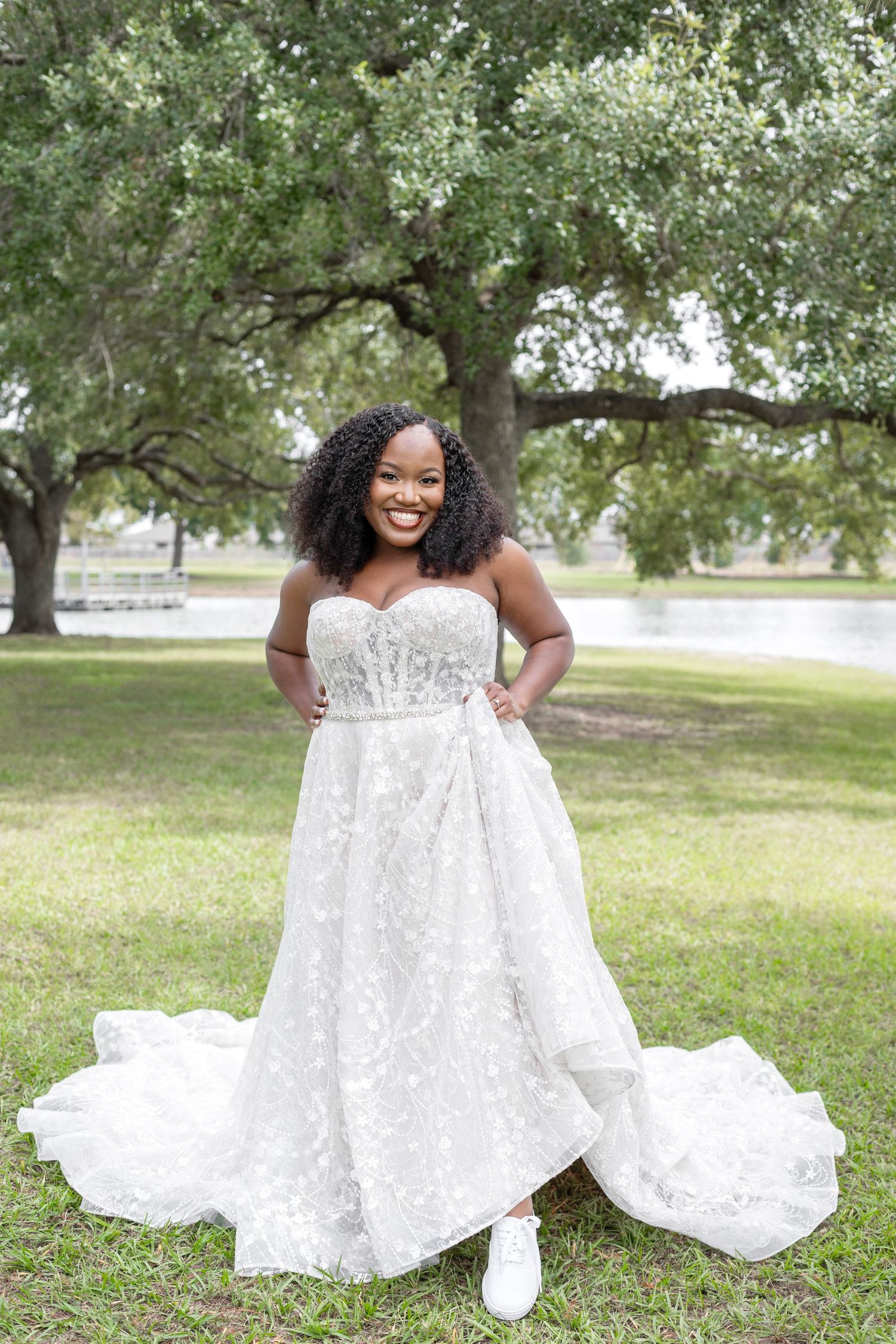 A bride in a stunning white lace dress poses playfully on green grass under trees.