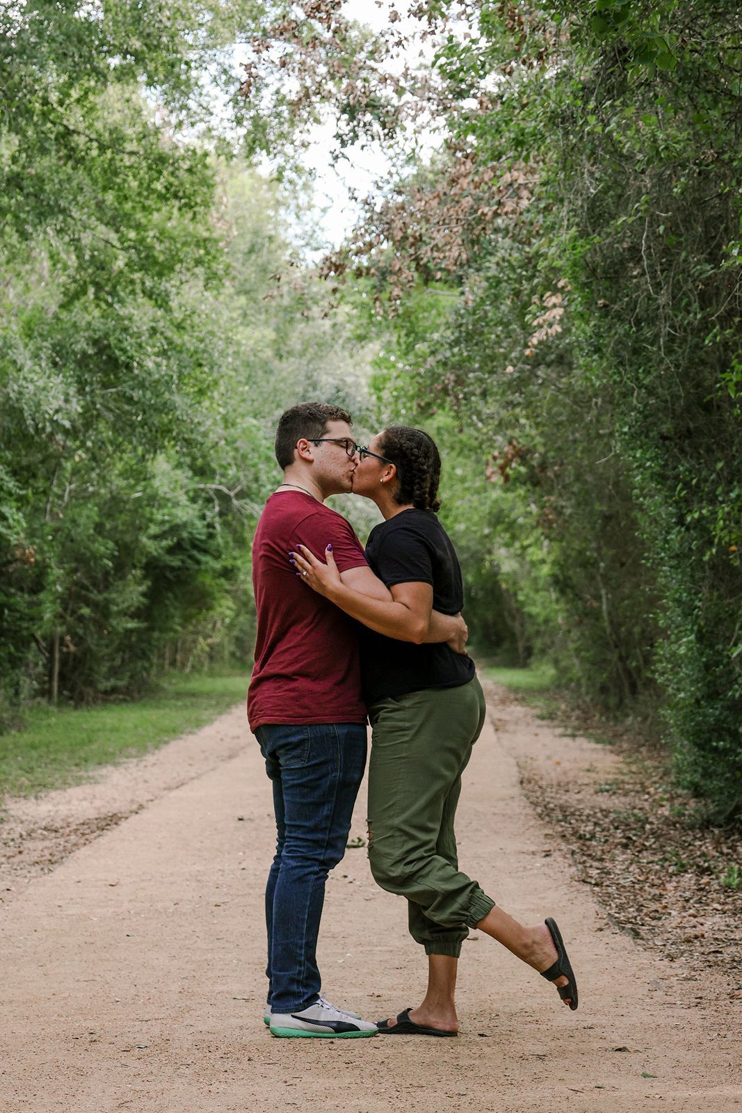 Couple sharing a kiss on a scenic dirt path surrounded by lush green trees in a romantic outdoor setting.