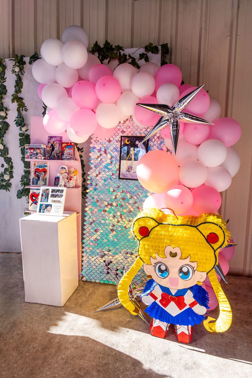 Sailor Moon piñata at a party with pink and white balloon arch, sequin backdrop, and ivy decorations.